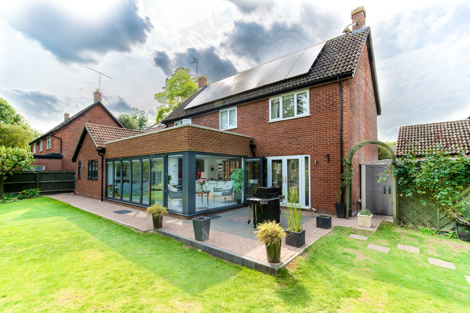 Exterior of a modern brick home with a new orangery kitchen extension featuring dark-framed bifold doors opening onto a patio.